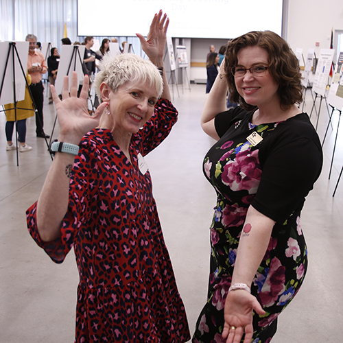 Two women stand sideways to the camera with their arms open in a presentational gesture.