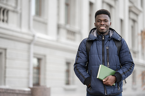 Black, male student smiling at the camera wearing a coat and holding book