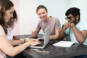 "A diverse group of students sit around a table and work together on a laptop computer."