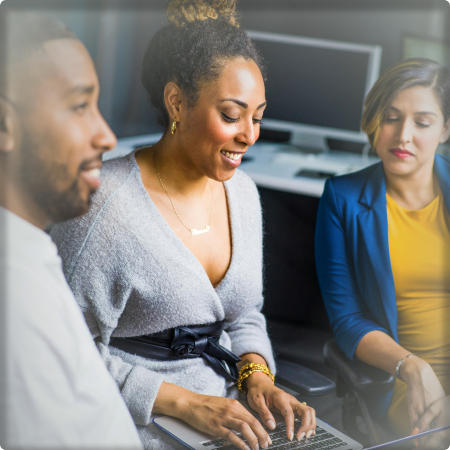 A photo of three people, professionally dressed, the person in the center is typing on a laptop.