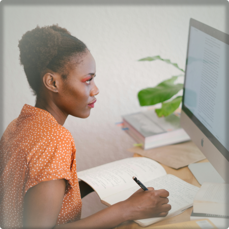 A photo of a woman writing in a notebook in front of a computer. Taken from the right.