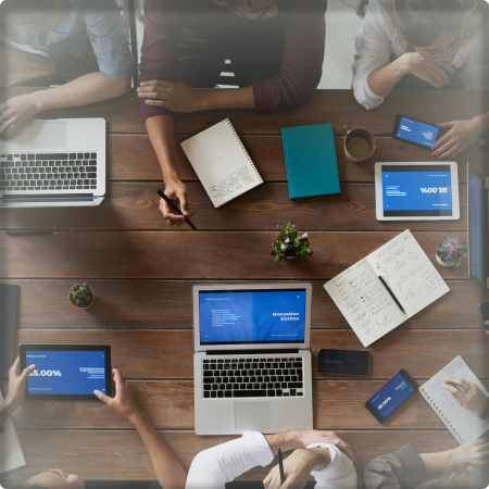 An overhead image of a table with seven people sitting at it. There is one person sitting at the head of the table and the other six members of the photo are looking to them with various notetaking devices in front of them (notebooks, laptops, tablets, etc.).